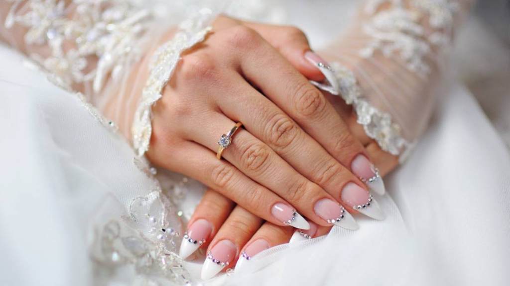 Bride holding bouquet with elegant oval nude wedding nails glistening in sunlight.